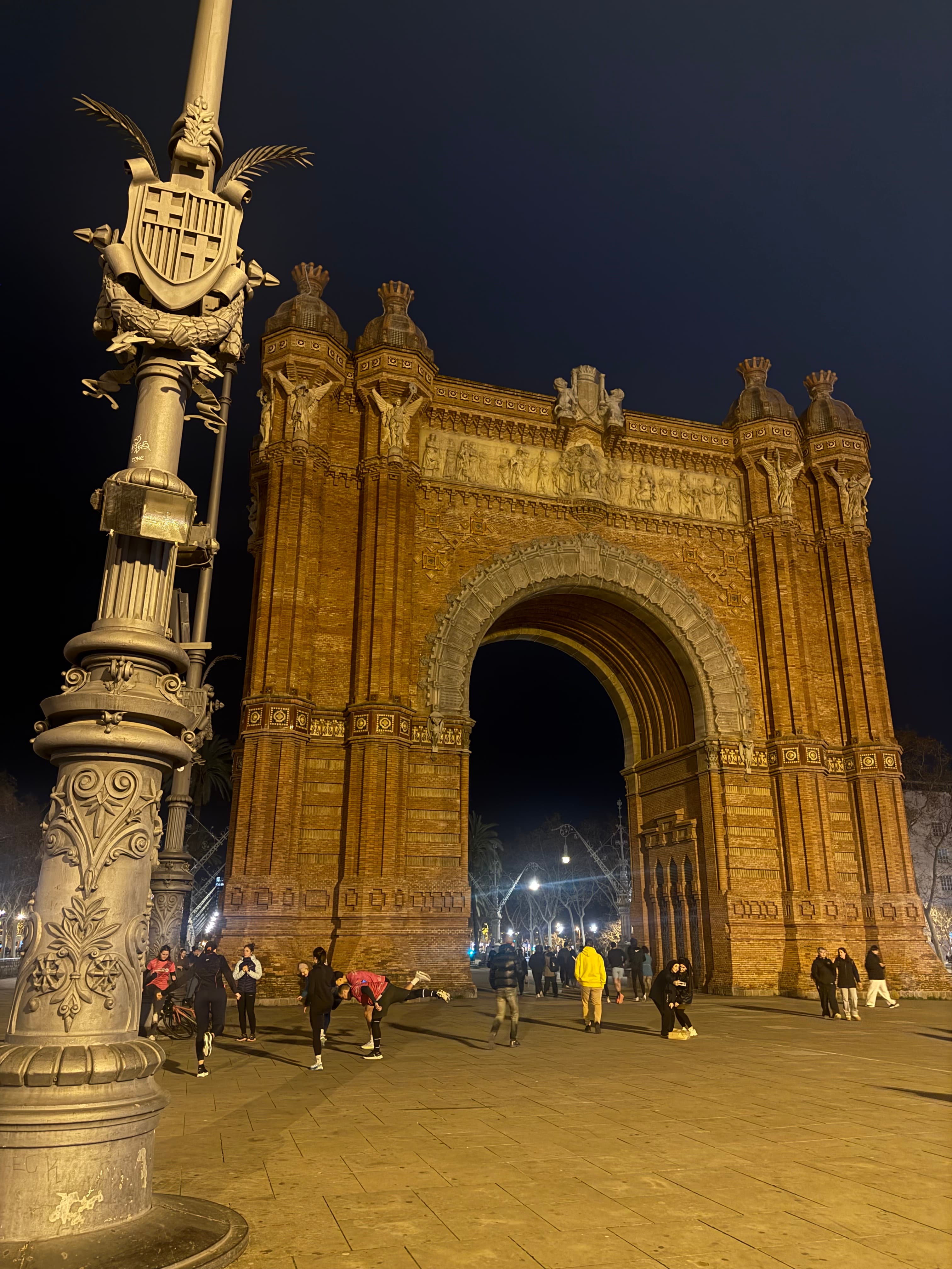 Barcelona's red brick Arc de Triomf at night with people and an ornate lamppost.